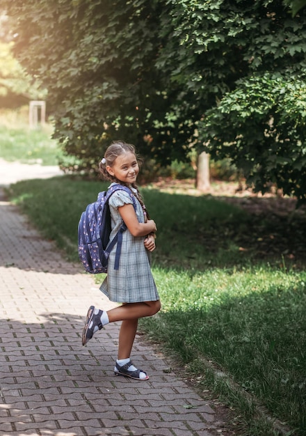 A little girl in a dress with pigtails holds large blue backpack and walks on road alley First september day in elementary school