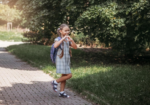 A little girl in a dress with pigtails holds large blue backpack and walks on road alley First september day in elementary school