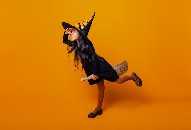 Little girl dressed as a Halloween witch in a black dress and hat flies on a broomstick on a yellow background.