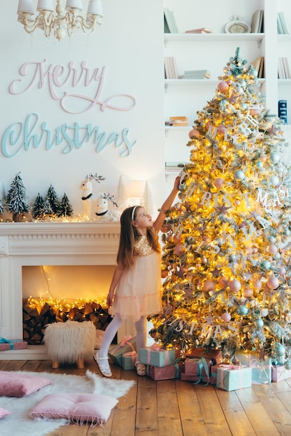 little girl, dressed in a beautiful fashionable white dress, poses near the Christmas tree.