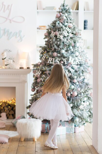 little girl, dressed in a beautiful fashionable white dress, poses near the Christmas tree.