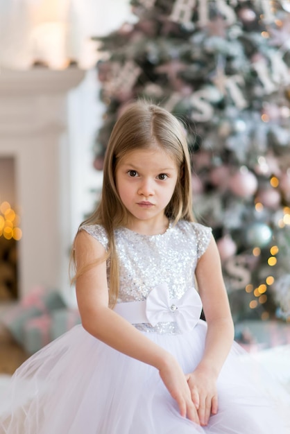 little girl, dressed in a beautiful fashionable white dress, poses near the Christmas tree.