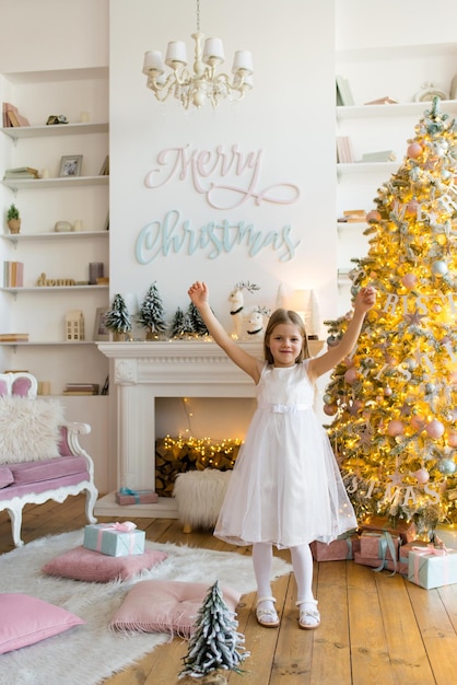 little girl, dressed in a beautiful fashionable white dress, poses near the Christmas tree.