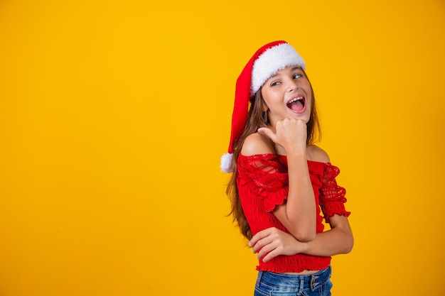 Little girl dressed for christmas pointing with her thumb on free space for text. Year-end promotion and campaigns