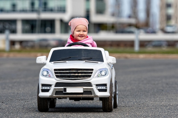 Little girl drives a big white childrens electric toy car in the parking lot at the residential home