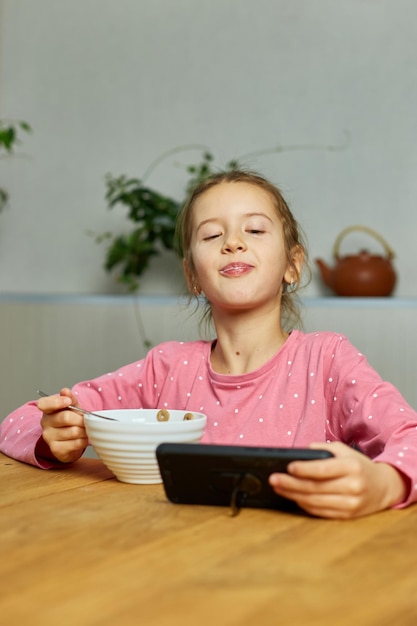 Little girl eating cereal with milk and watching video on smartphone