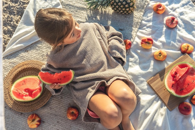 Little girl eats fruit lying on a blanket on the beach