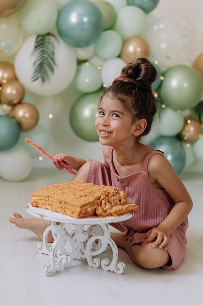 Little girl eats a square layer cake in a room decorated with balloons the concept of a birthday