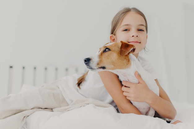 Little girl embraces small pedigree dog stay in bed plays with favourite pet before sleep has appealing look gazes camly into camera Childhood and bed time concept Kid hugs domestic animal