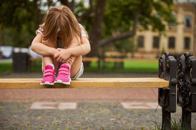 Little girl feeling bored while sitting on the wooden bench in the city park