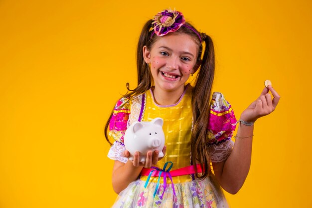 Little girl in festa junina clothes holding a piggy bank on yellow background Finance and festa junina concept