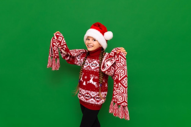 A little girl in a festive Christmas sweater and scarf on a green isolated background is very happy about the holiday