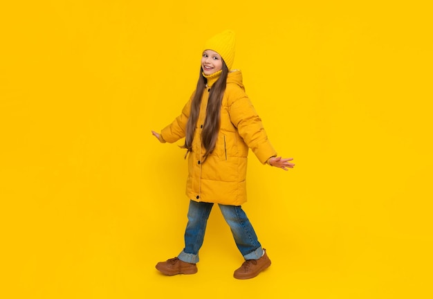 Photo a little girl in full height in an autumn coat down jackets and hats rejoices in autumn autumn clothes for children yellow isolated background