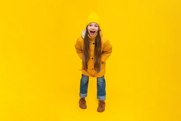 Photo a little girl in full height in a down jacket and a hat smiles with her mouth wide open the child is happy about the onset of autumn warm clothes for children yellow isolated background