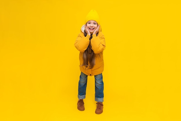 A little girl in full height in a warm jacket and hat rejoices at the onset of autumn Beautiful young happy girl on a yellow isolated background in the studio