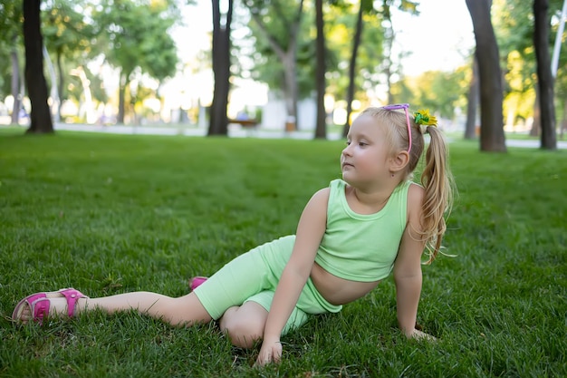 A little girl in a green suit sits on the lawn in the park and poses for the camera