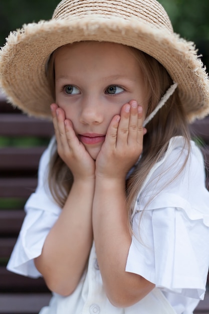 A little girl in a hat looks at the camera with her hand held to her face