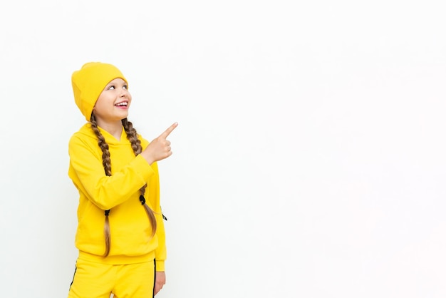 A little girl in a hat and a yellow suit points to your advertisement on a white isolated background Autumn clothes for children Copy space