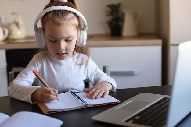 Little girl in headphones sit at desk writing in notebook studying online do exercises at home, little child handwrite prepare homework on quarantine, have web class or lesson indoors.