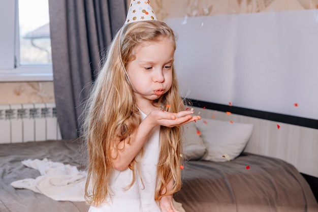 Little girl on her birthday morning inflates multicolored confetti from palm her hand in festive cap