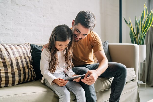 Little girl and her father having fun together while playing with a digital tablet at home. Monoparental family concept.