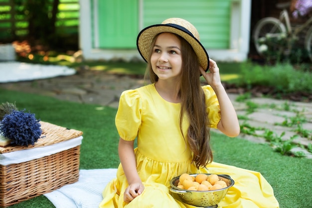 Little girl hold hat with hand sit with apricots in a colander at picnic on lawn at summer