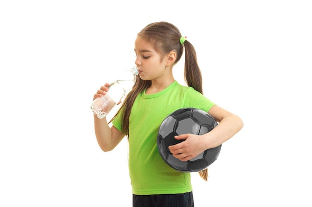 Little girl holding a ball in her hand and drinks water from a bottle is isolated on a white background