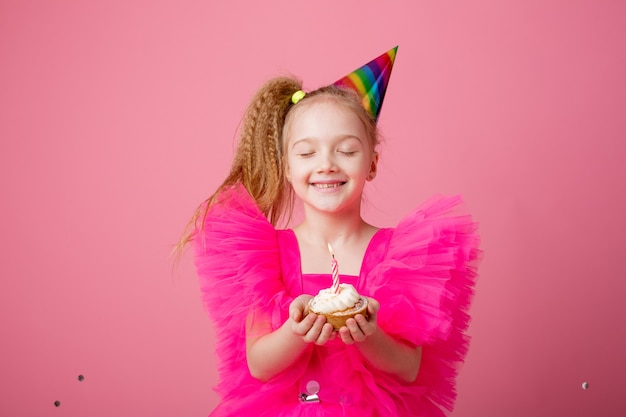 Little girl holding a festive cake blowing a candle on a pink background celebrating her birthday