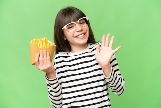 Little girl holding fried chips over isolated chroma key background saluting with hand with happy expression