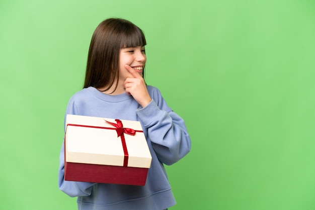 Little girl holding a gift over isolated chroma key background thinking an idea and looking side