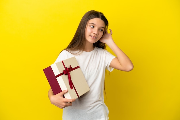 Little girl holding a gift over isolated yellow background listening to something by putting hand on the ear