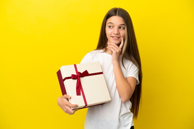 Little girl holding a gift over isolated yellow background thinking an idea while looking up