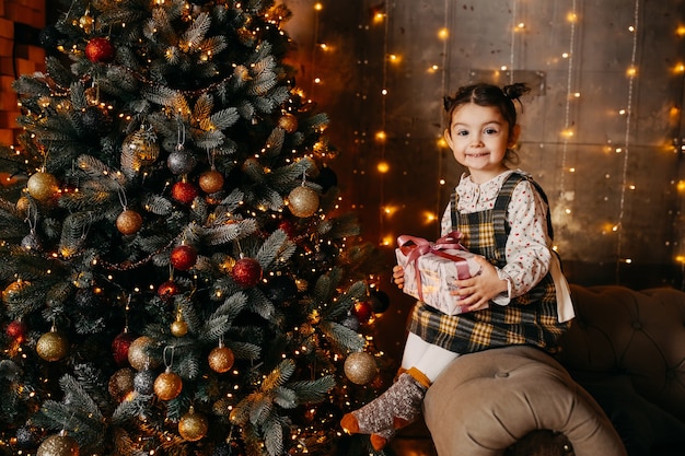 Little girl holding a gift, sitting next to Christmas tree