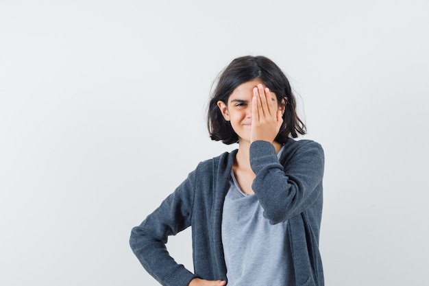 Little girl holding hand on eye in t-shirt, jacket and looking merry. front view.