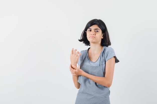 Little girl holding hand on her wrist in t-shirt, apron and looking confused , front view.