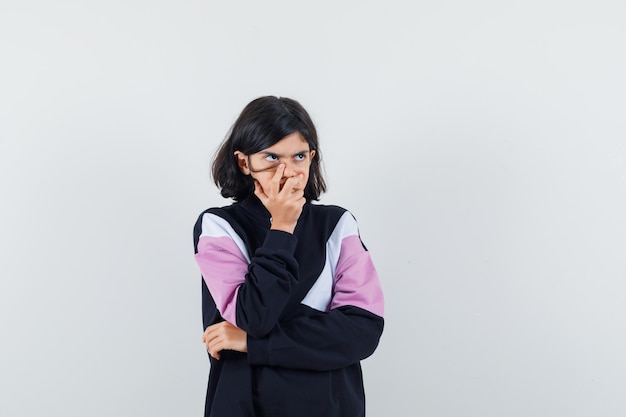 Little girl holding hand on mouth in shirt and looking pensive , front view.