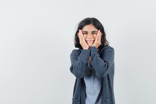 Little girl holding hands on cheeks in t-shirt, jacket and looking cheery. front view.