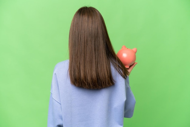 Little girl holding a piggybank over isolated chroma key background in back position