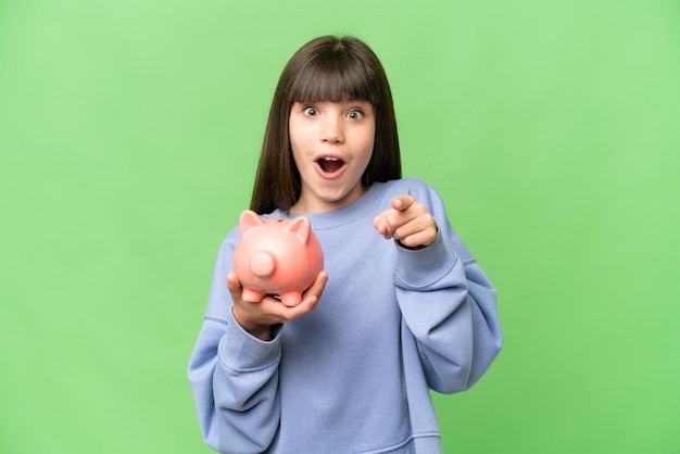 Little girl holding a piggybank over isolated chroma key background surprised and pointing front