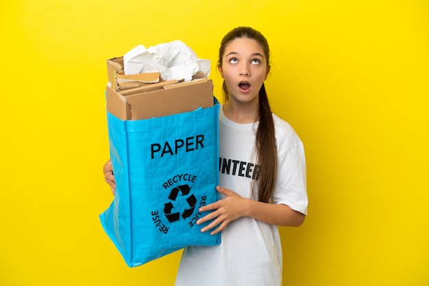 Little girl holding a recycling bag full of paper to recycle over isolated yellow background looking up and with surprised expression