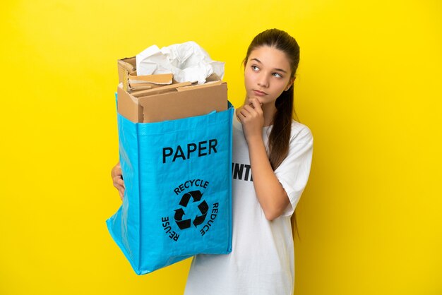 Little girl holding a recycling bag full of paper to recycle over isolated yellow background and looking up