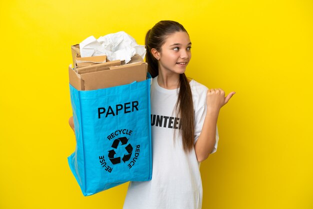 Little girl holding a recycling bag full of paper to recycle over isolated yellow background pointing to the side to present a product