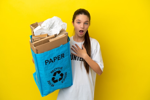 Little girl holding a recycling bag full of paper to recycle over isolated yellow background surprised and shocked while looking right