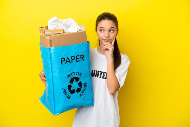 Little girl holding a recycling bag full of paper to recycle over isolated yellow background thinking an idea while looking up