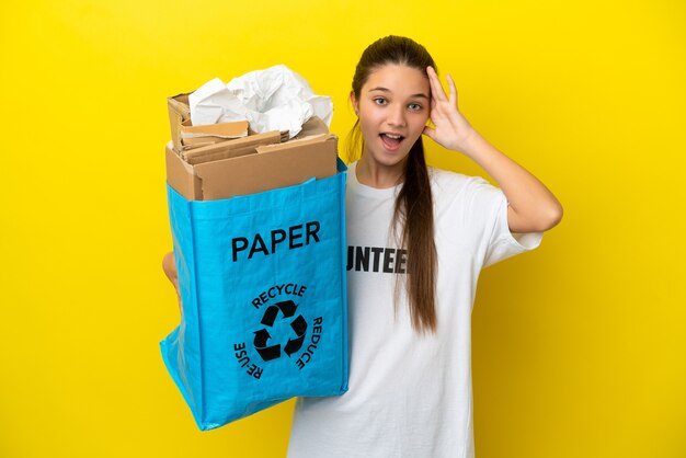 Little girl holding a recycling bag full of paper to recycle over isolated yellow background with surprise expression