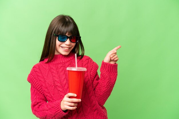 Little girl holding soda over isolated chroma key background pointing finger to the side