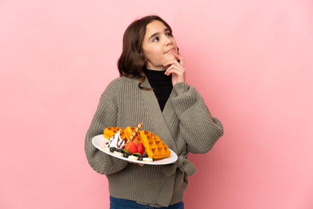 Little girl holding waffles isolated on pink background having doubts while looking up