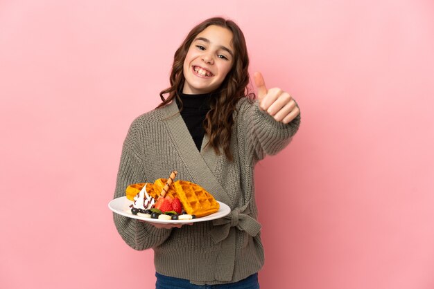 Little girl holding waffles isolated on pink background with thumbs up because something good has happened