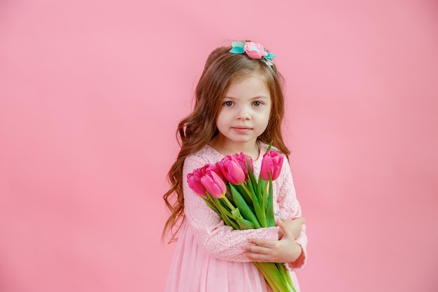 A little girl holds a bouquet of pink tulips on a pink background