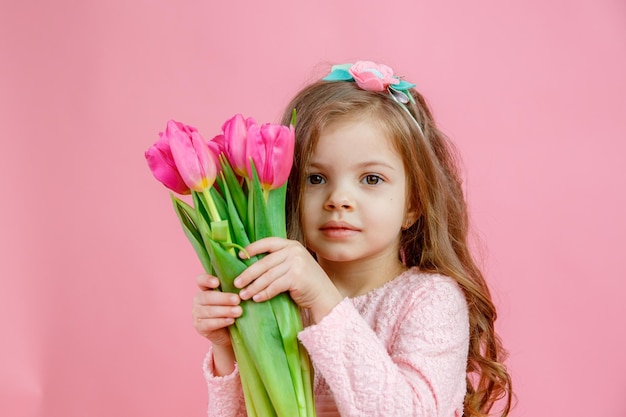 A little girl holds a bouquet of pink tulips on a pink background